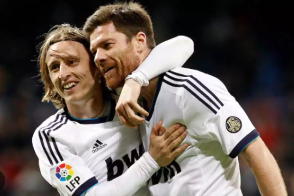 Two players in white uniforms celebrate a goal by hugging on the field.