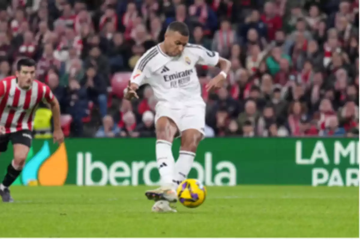 Soccer player in a white uniform kicking a ball during a match while an opposing player in a red and white striped uniform watches him.