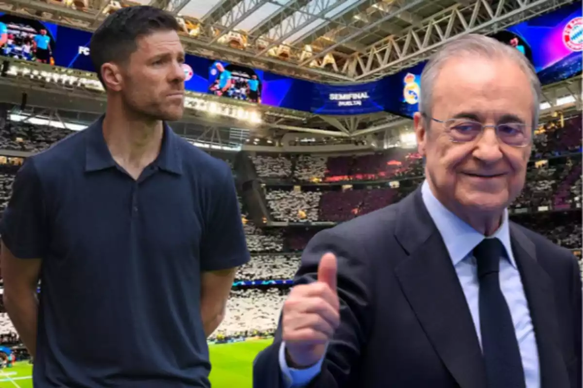 Two men in a packed soccer stadium, one wearing a blue polo jersey and the other in a suit and tie giving a thumbs-up gesture