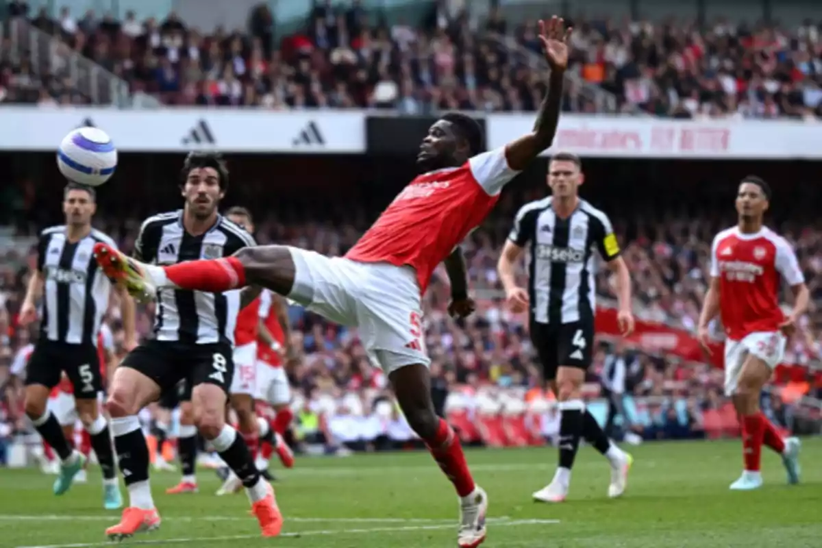 A player in a red and white uniform tries to control the ball in the air during a match against a team in black and white striped uniforms in a stadium full of spectators.