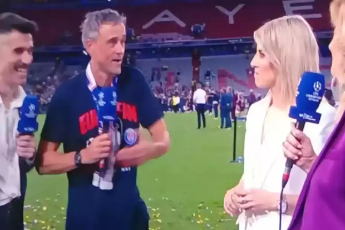 Three people with blue microphones talk in a soccer stadium after a match, with spectators and confetti on the field.