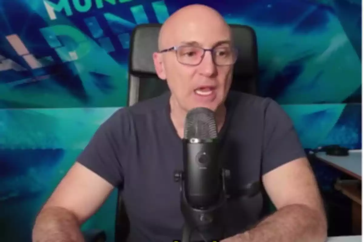Bald man with glasses speaking in front of a microphone in a studio with a blue background