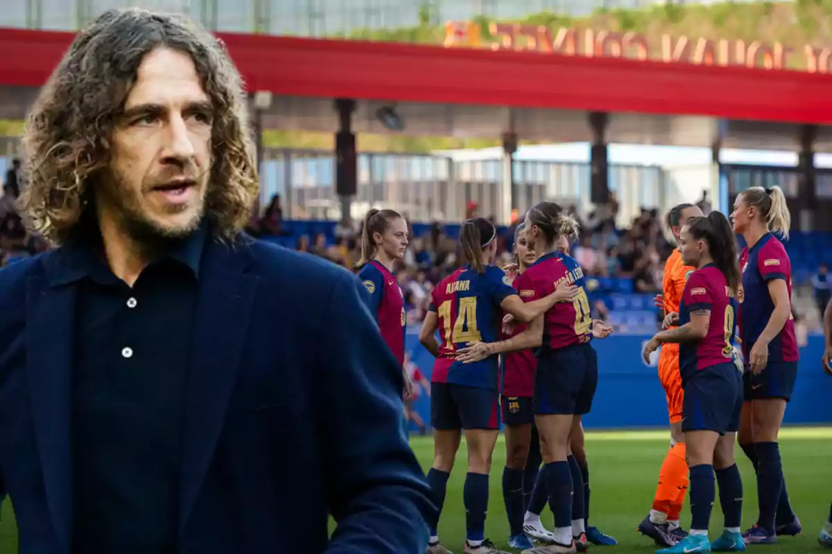 A man with curly hair and a dark jacket appears in the foreground while a group of female soccer players embrace on the field in the background.