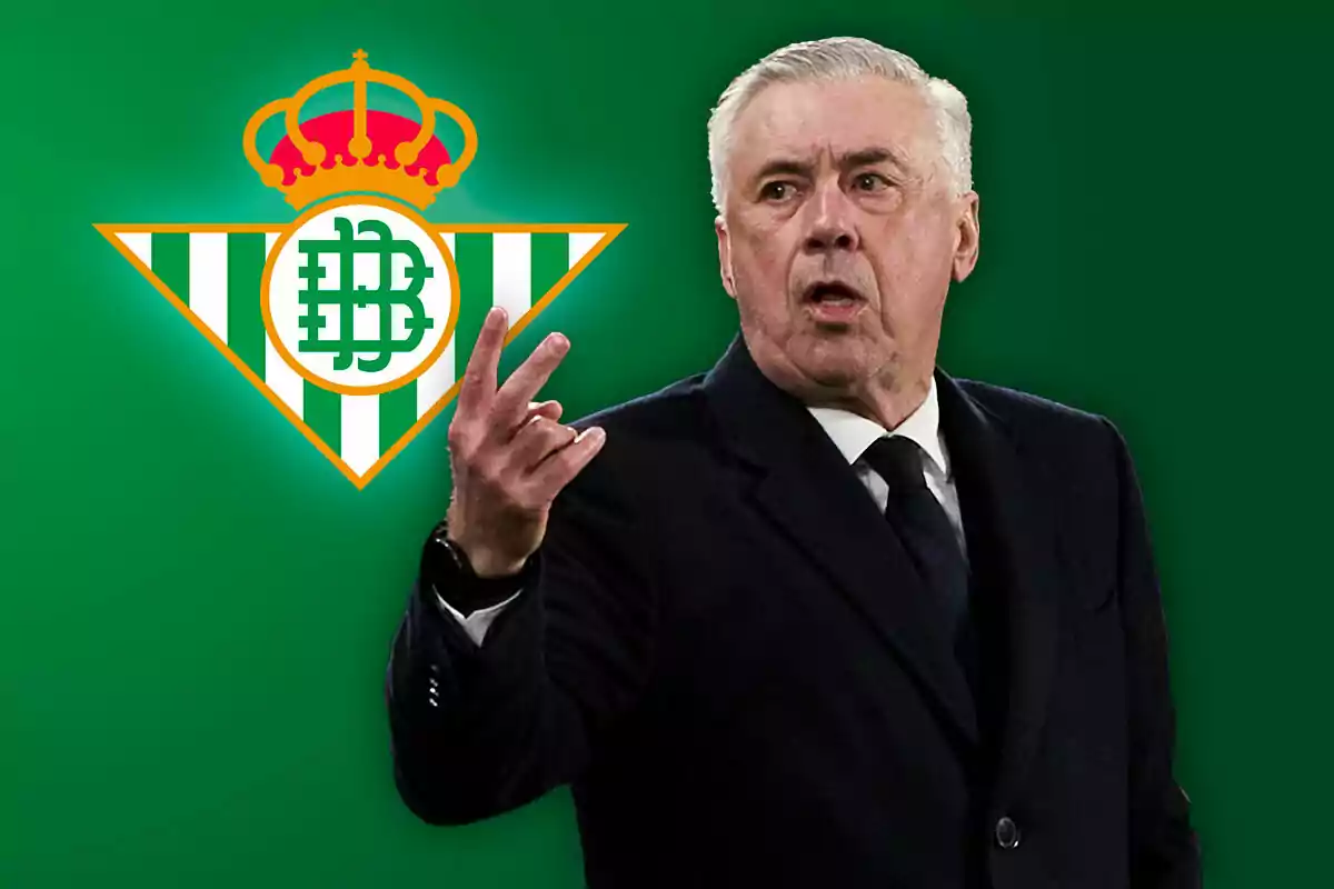 Un hombre de traje gesticulando con la mano frente al logo del Real Betis en un fondo verde.