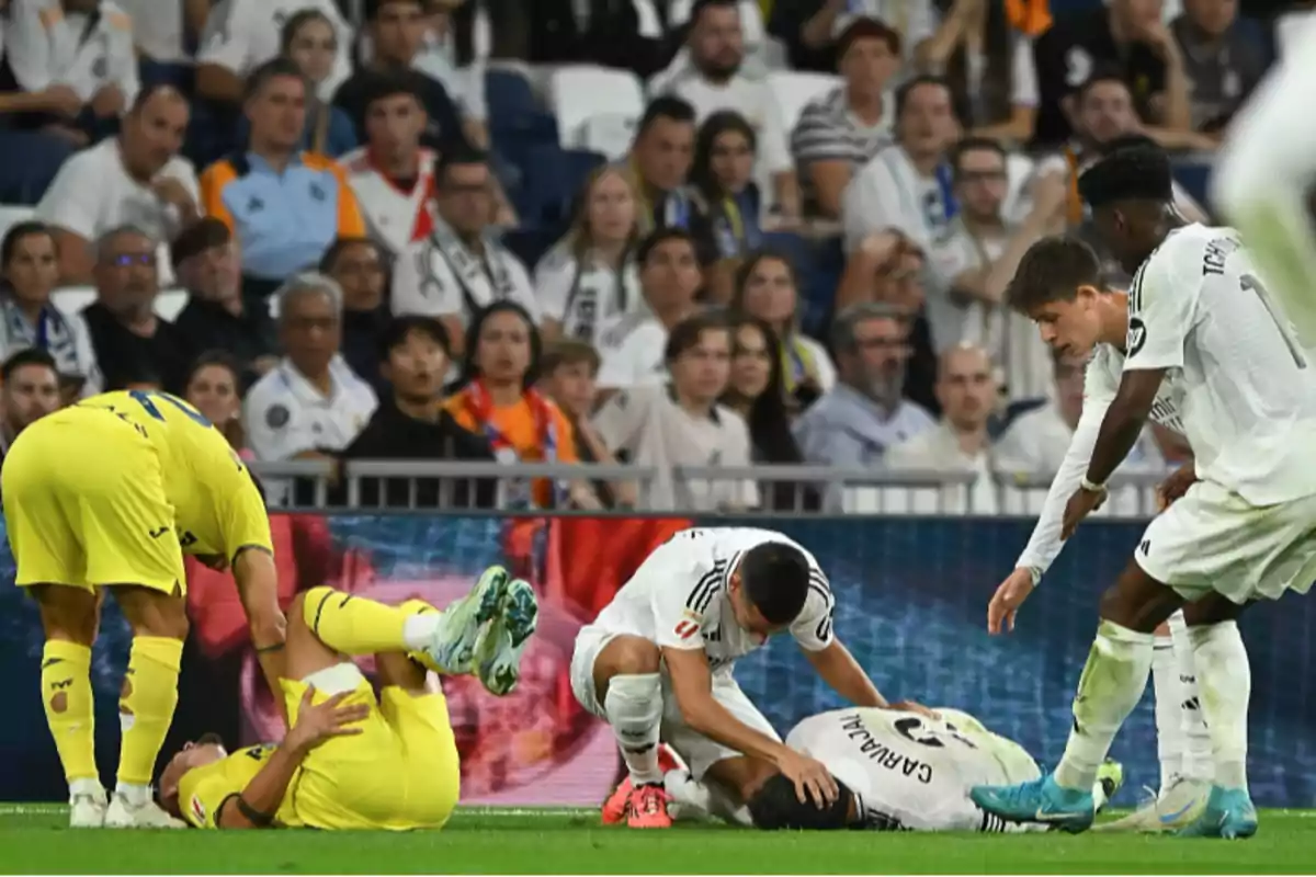 Soccer players on the field, one of them on the ground while others assist him, with spectators in the background.