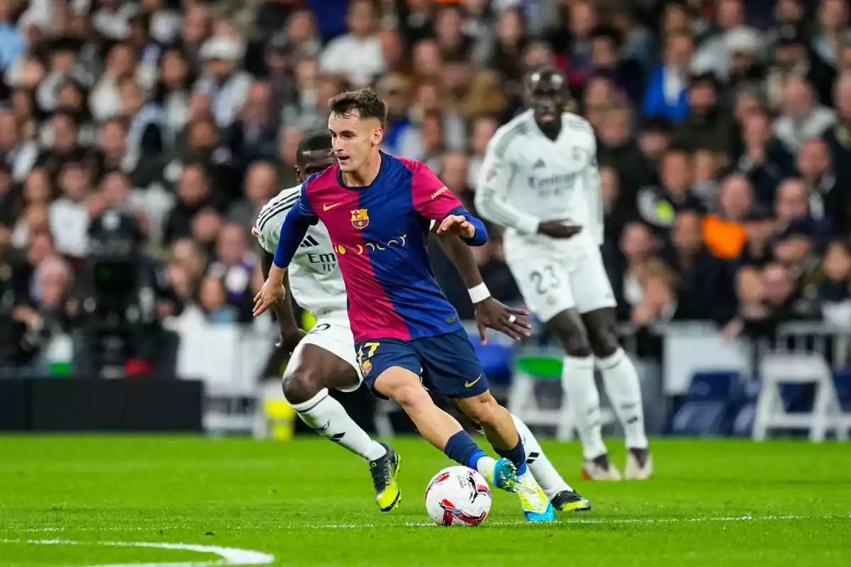 Un jugador de fútbol con uniforme azul y rojo controla el balón mientras es perseguido por jugadores con uniforme blanco en un estadio lleno de espectadores.