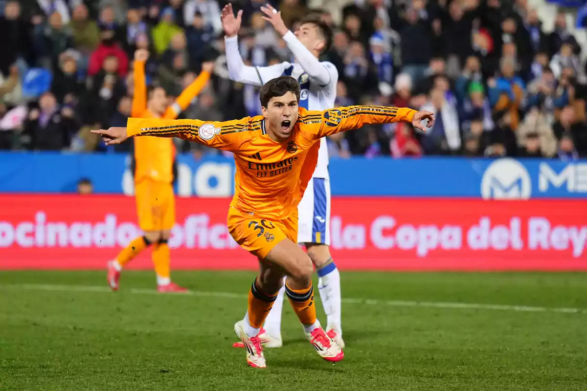 A player in an orange uniform celebrates enthusiastically on the field while another player in the background raises his arms.