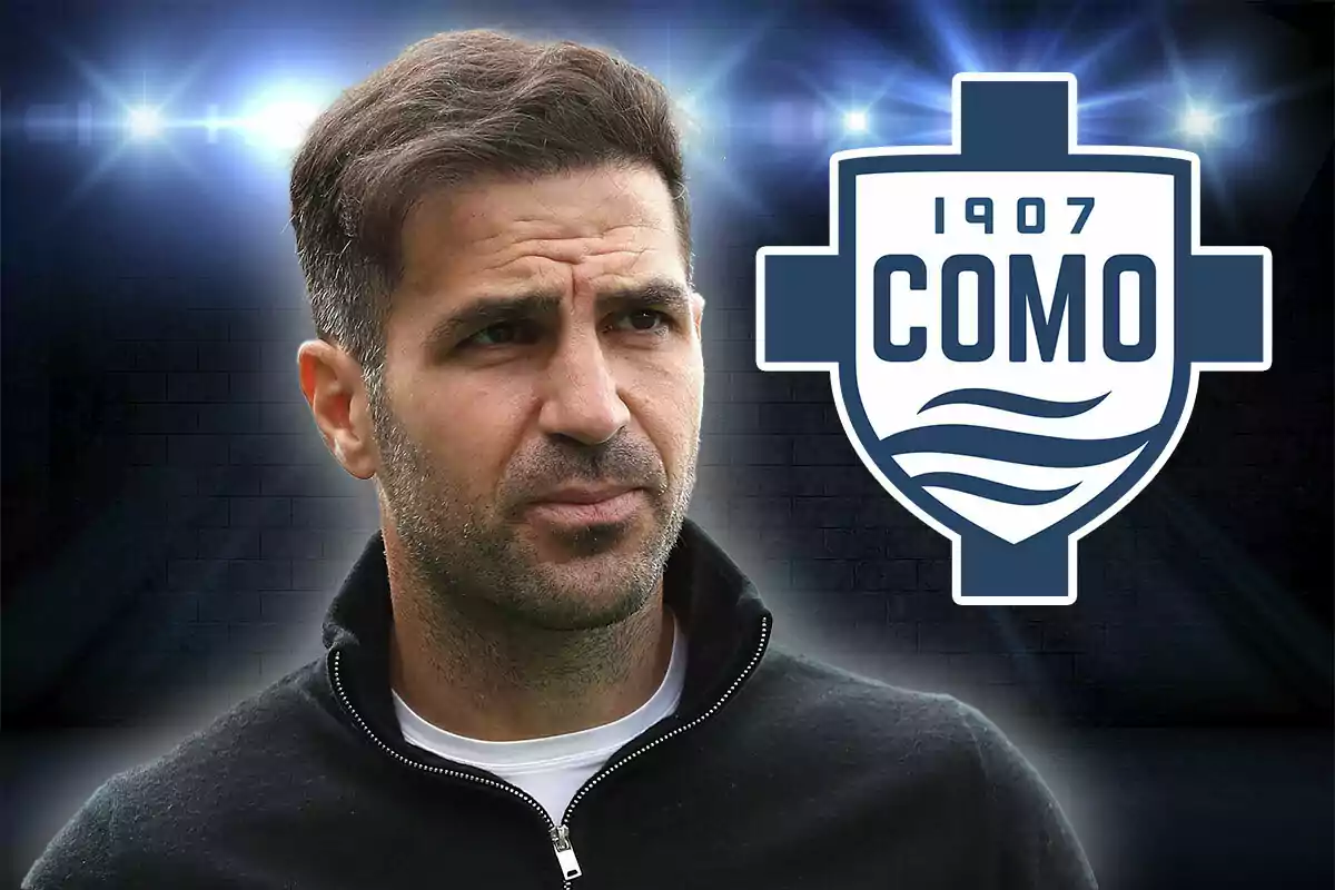 Un hombre con barba y cabello oscuro mira hacia un lado con una expresión seria mientras lleva una chaqueta negra y una camiseta blanca con el logotipo del equipo de fútbol Como 1907 al fondo sobre un fondo oscuro iluminado por luces brillantes.