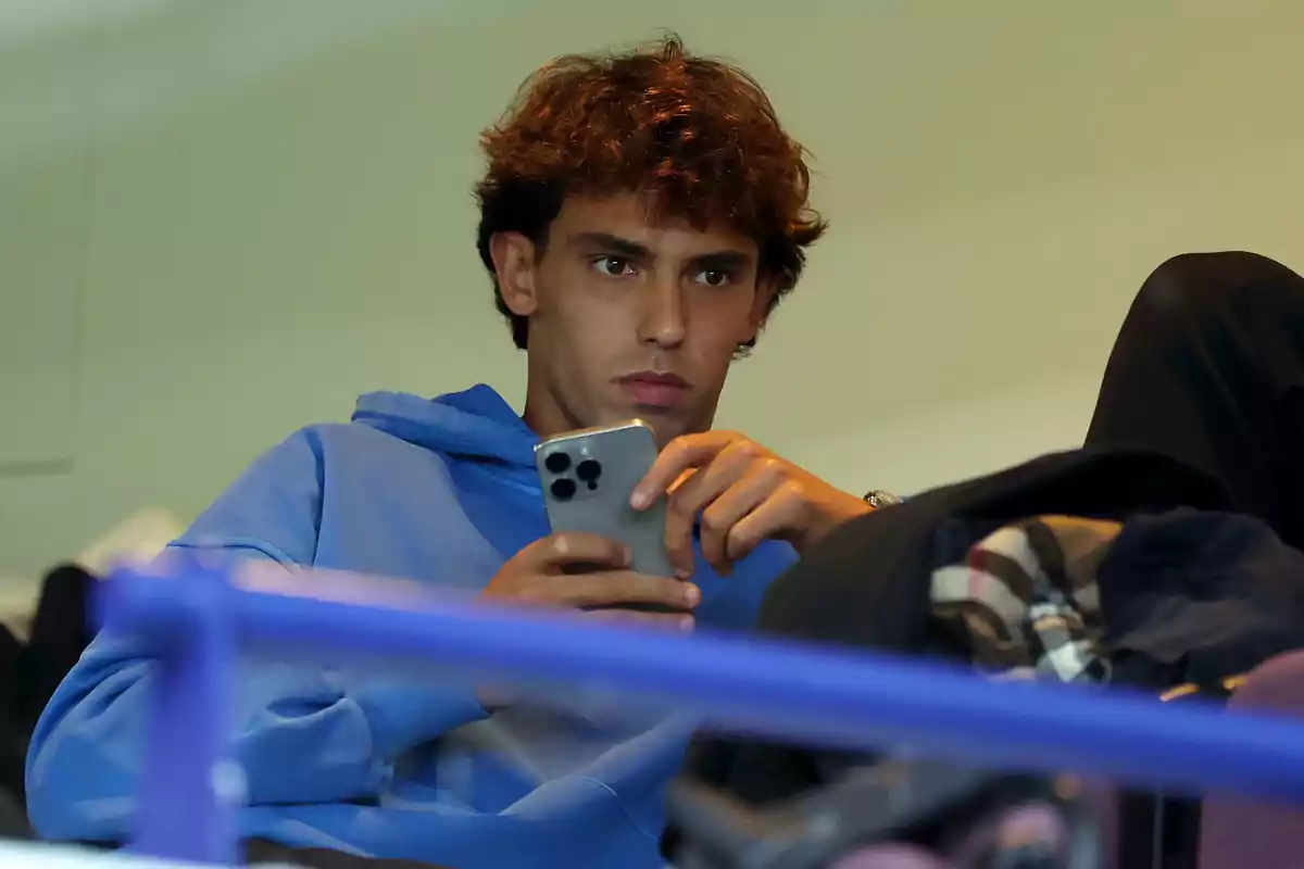 A young man in a blue sweatjersey holds a mobile phone while sitting.