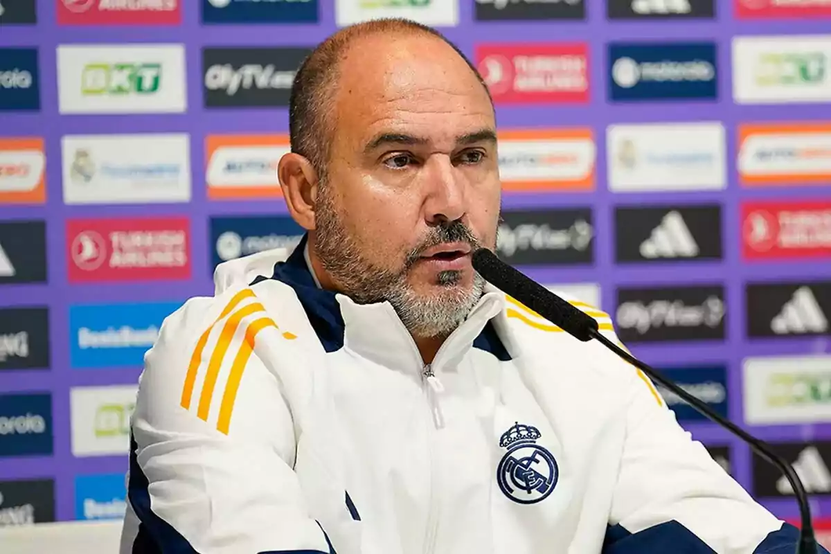 Chus Mateo at a press conference wearing a Real Madrid tracksuit and a microphone in front of him.