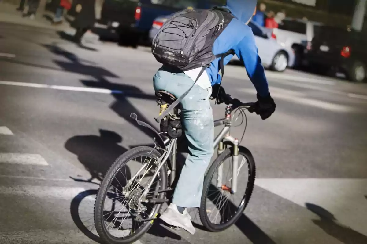 Persona con sudadera azul y mochila negra montando una bicicleta en una calle concurrida.