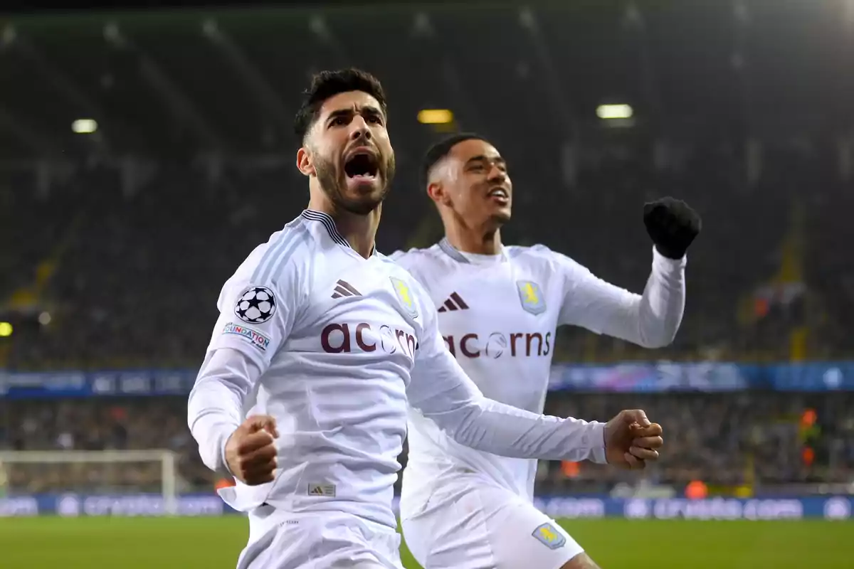 Two players celebrate a goal in a packed stadium while wearing Aston Villa’s white uniforms. Two players celebrate a goal in a packed stadium while wearing Aston Villa’s white uniforms.