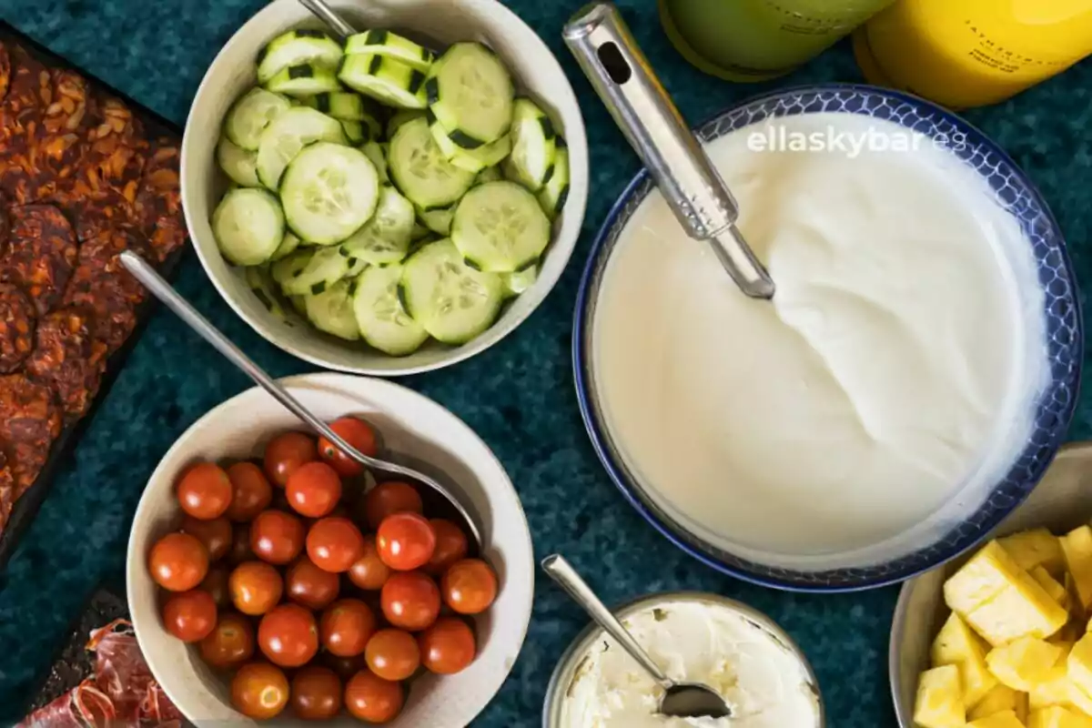 Una mesa con varios platos que contienen pepinos en rodajas, tomates cherry, piña en trozos, una salsa blanca y un plato de comida cocinada. Una mesa con varios platos que contienen pepinos en rodajas, tomates cherry, piña en trozos, una salsa blanca y un plato de comida cocinada.