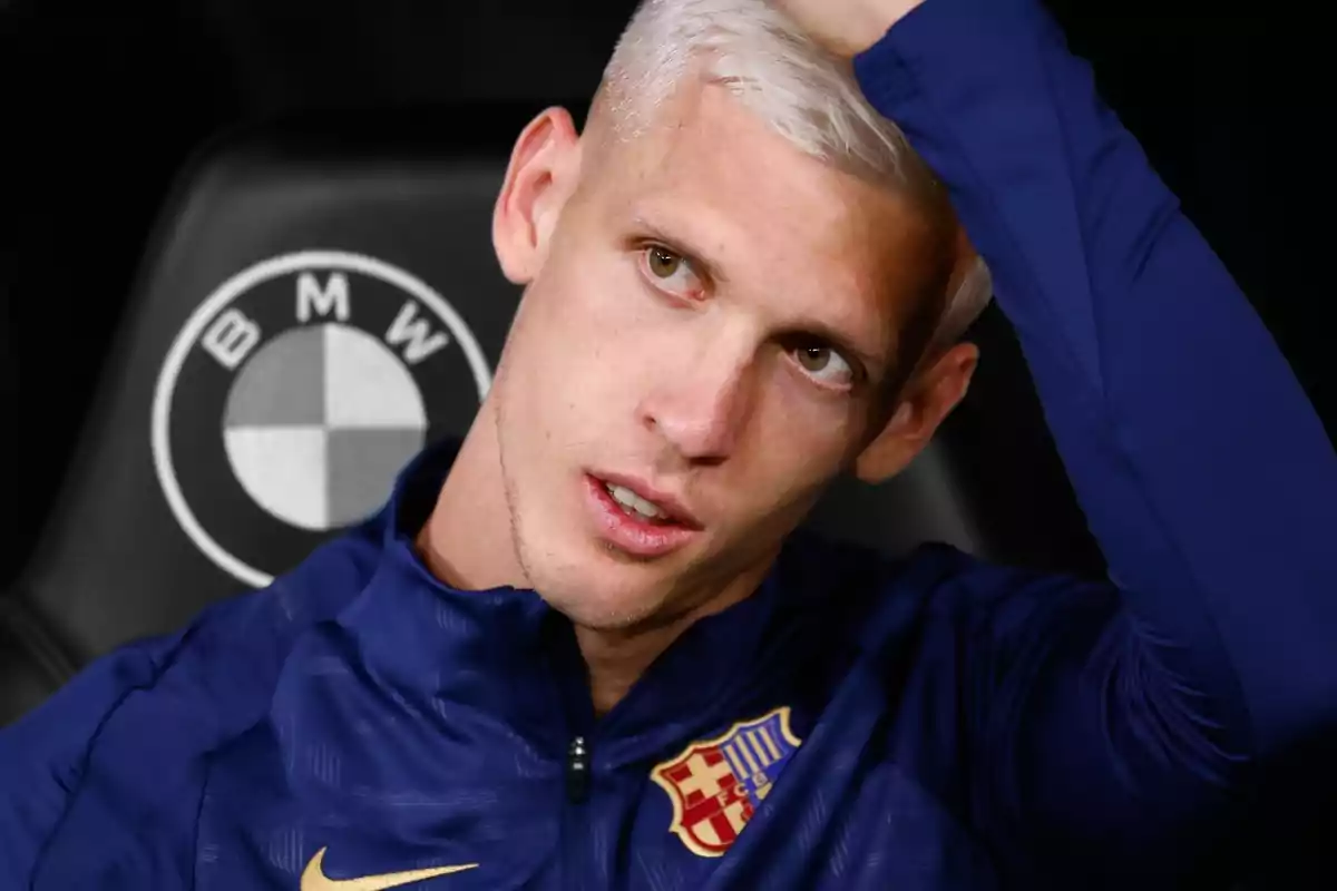 A soccer player with platinum blonde hair and a blue jacket with the FC Barcelona crest touches his head while sitting in front of a BMW logo.