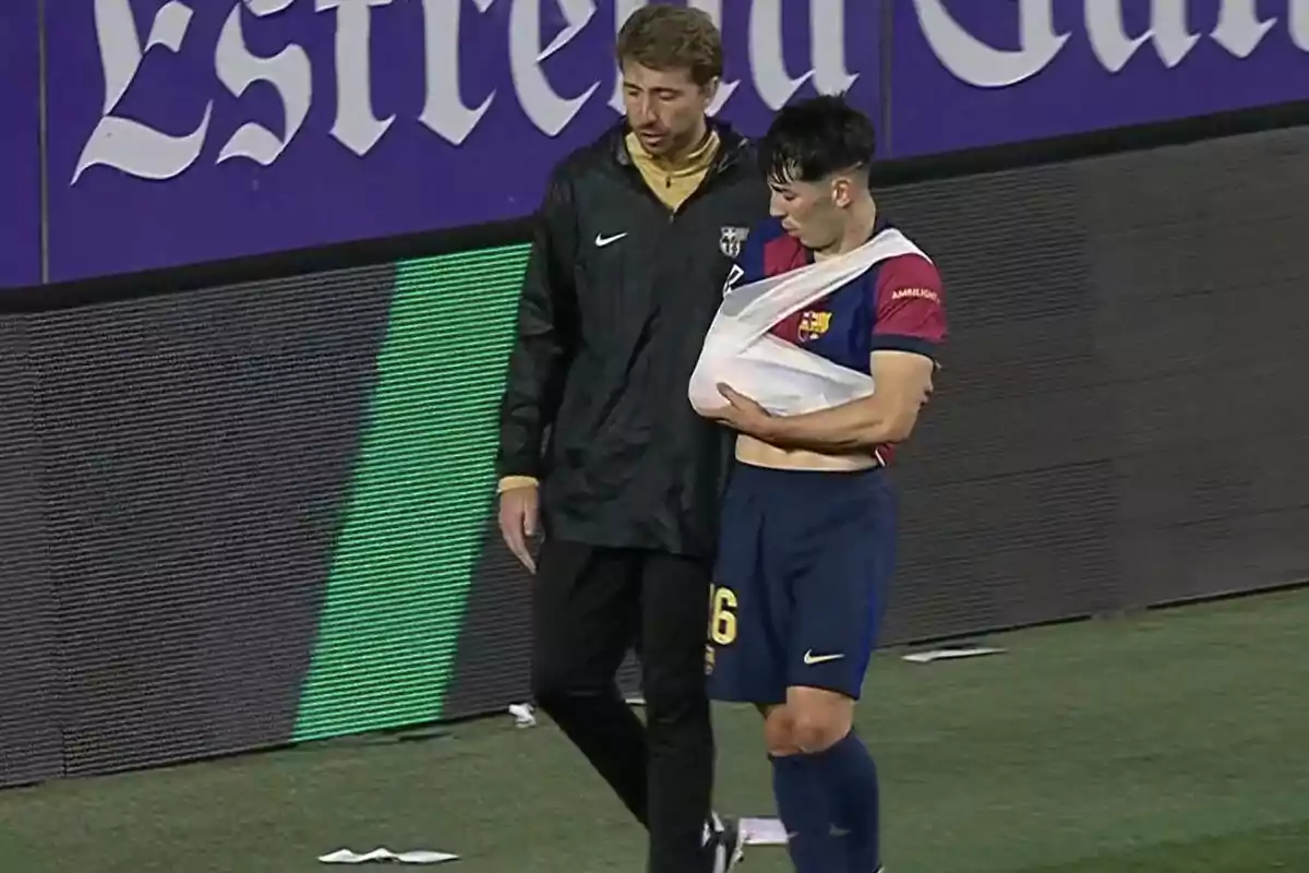 Un jugador de fútbol con el brazo en cabestrillo es acompañado por una persona del cuerpo técnico en el campo de juego. Un jugador de fútbol con el brazo en cabestrillo es acompañado por una persona del cuerpo técnico en el campo de juego.