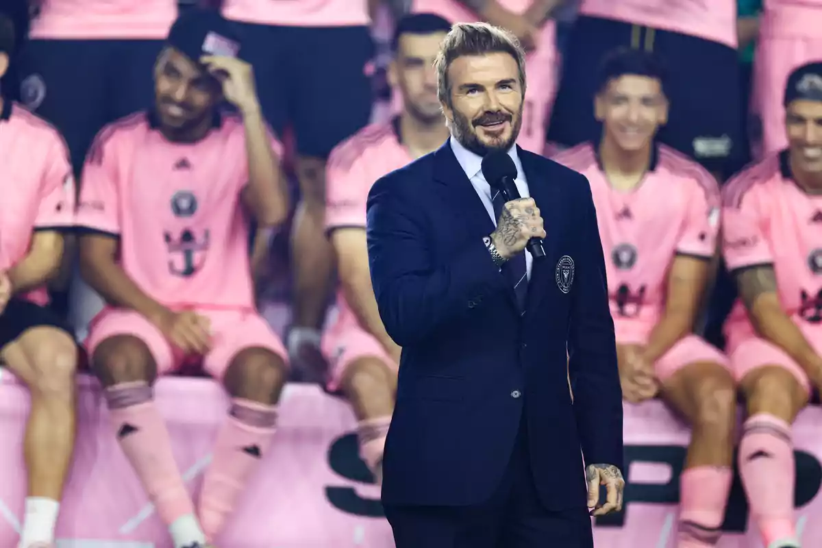 Un hombre con traje habla al micrófono frente a un grupo de jugadores de fútbol vestidos con uniformes rosas. Un hombre con traje habla al micrófono frente a un grupo de jugadores de fútbol vestidos con uniformes rosas.