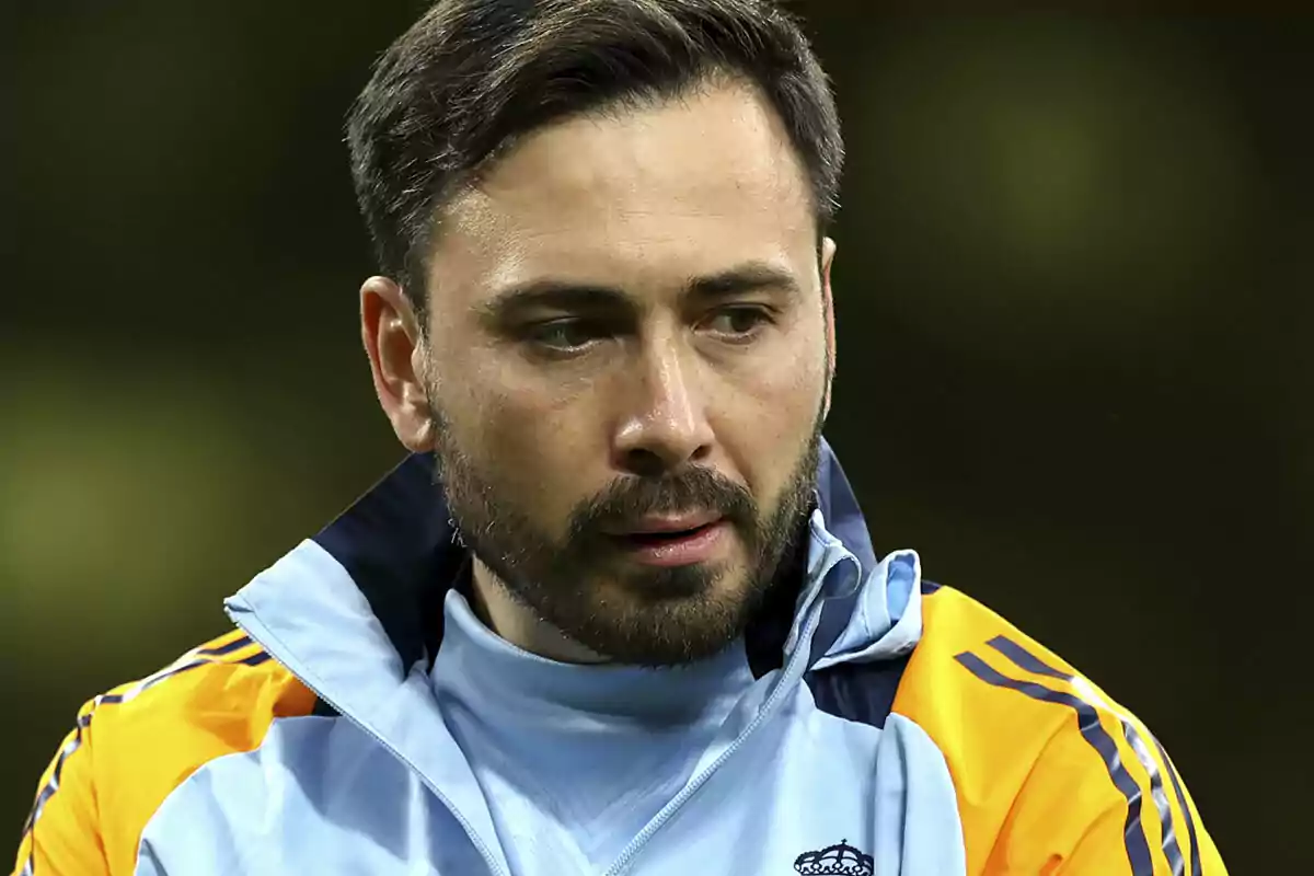 Hombre con barba y cabello corto vestido con chaqueta deportiva celeste y amarilla mirando hacia abajo