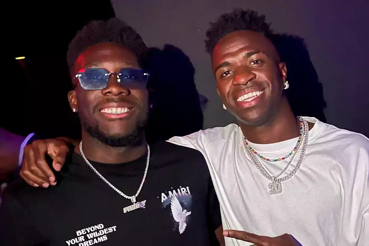 Davies y Vinicius sonrientes posando juntos, uno con gafas de sol y camiseta negra y el otro con camiseta blanca y collares.