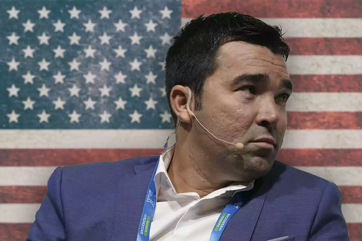 A man with a headset microphone, dressed in a blue suit, is sitting in front of a United States flag.