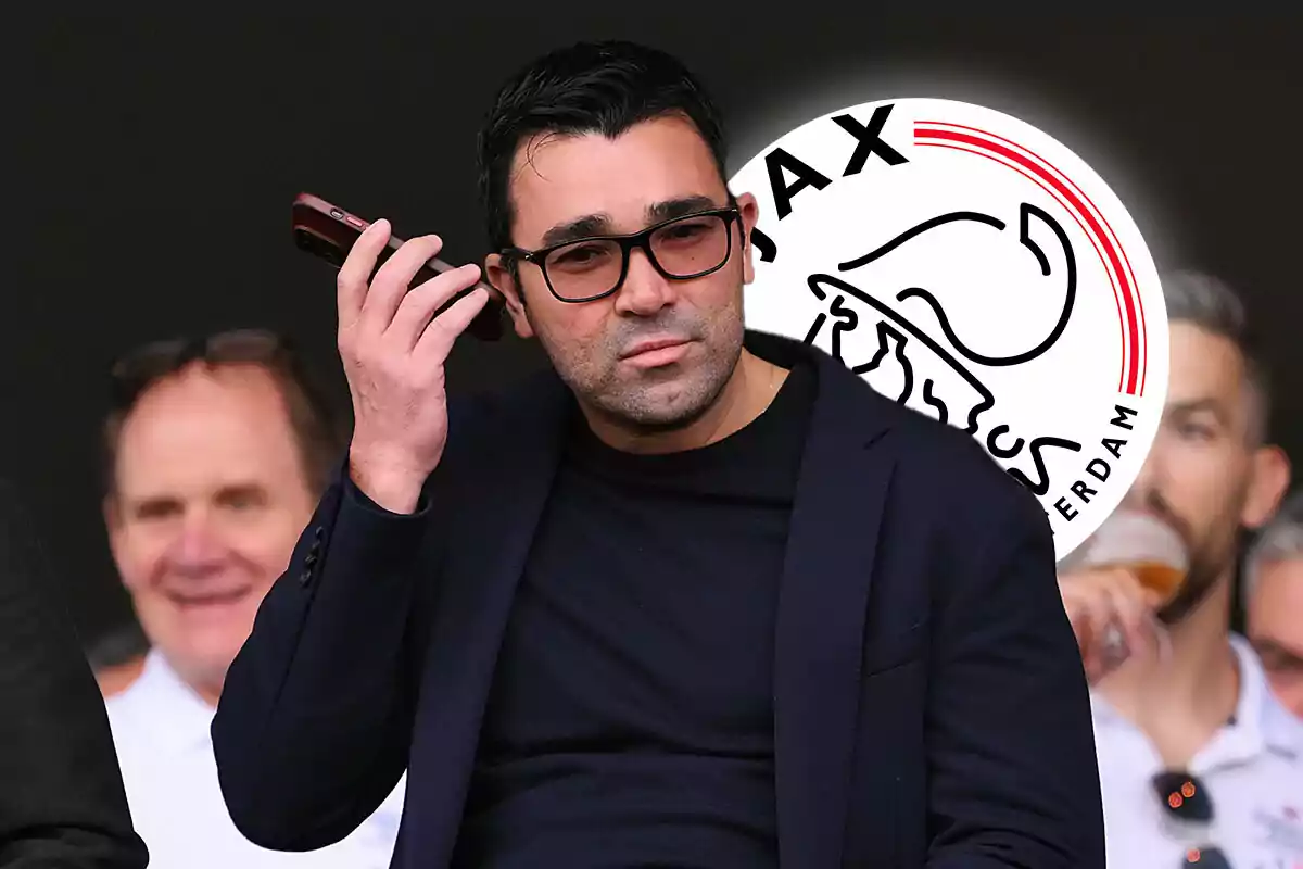 Deco con gafas y un teléfono en la mano, con el logo del Ajax de Ámsterdam de fondo.