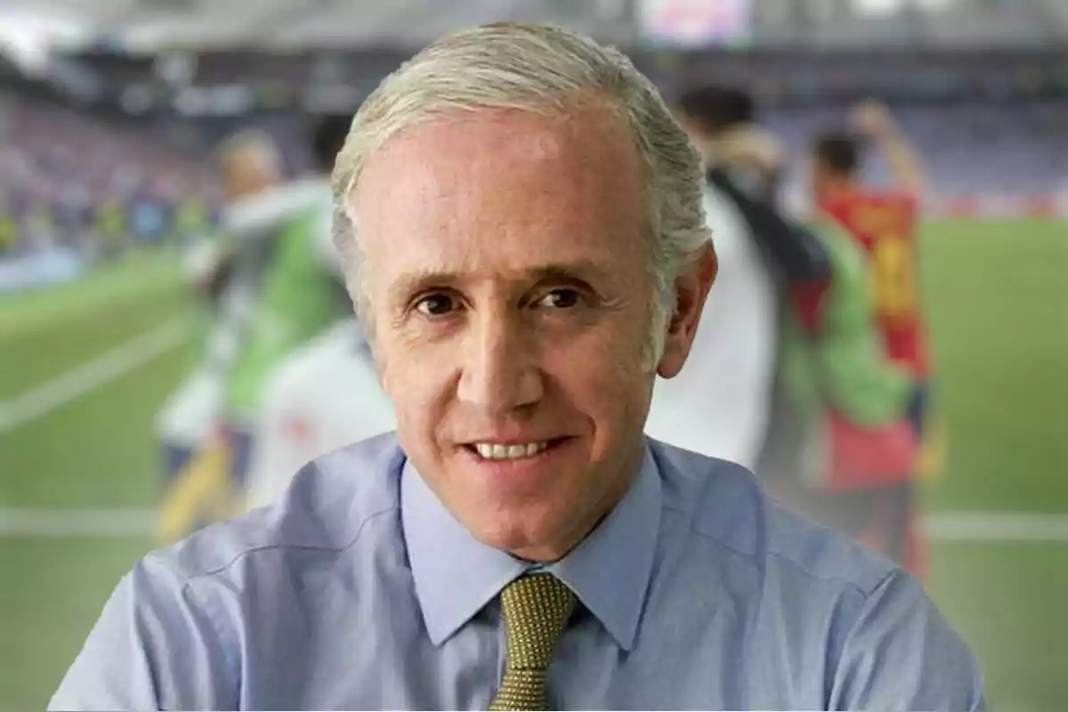 Eduardo Inda con camisa azul y corbata dorada sonriendo, con un estadio de fútbol desenfocado en el fondo.