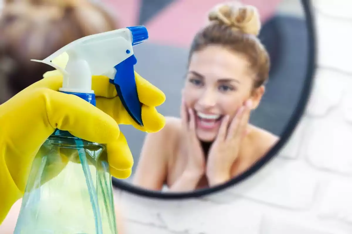 Una persona con guantes amarillos sostiene una botella de spray frente a un espejo en el que se refleja una mujer sonriente.