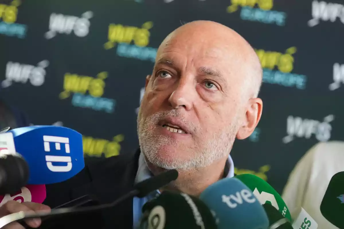 A bald man with a grey beard is speaking in front of several microphones from different media outlets, with a background that has the WFS Europe logo repeated.