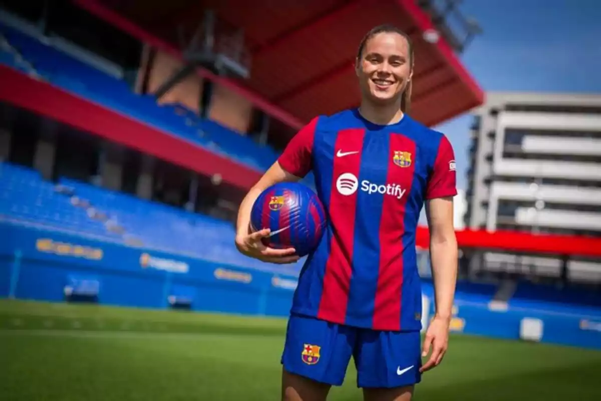 A female soccer player smiles as she holds a ball in a stadium with blue and red seats.