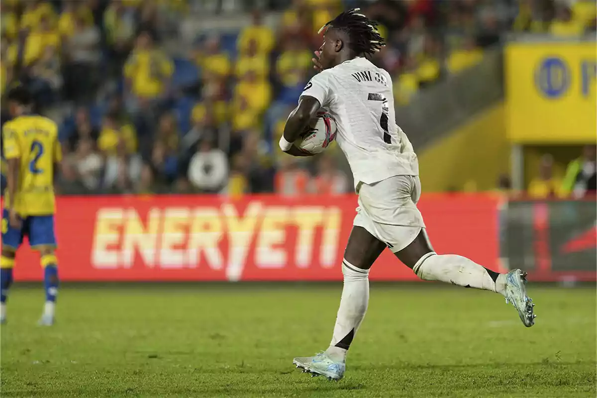 Vinicius con uniforme del Real Madrid corre sosteniendo el balón mientras el público y otros jugadores con uniforme de Las Palmas se ven al fondo.