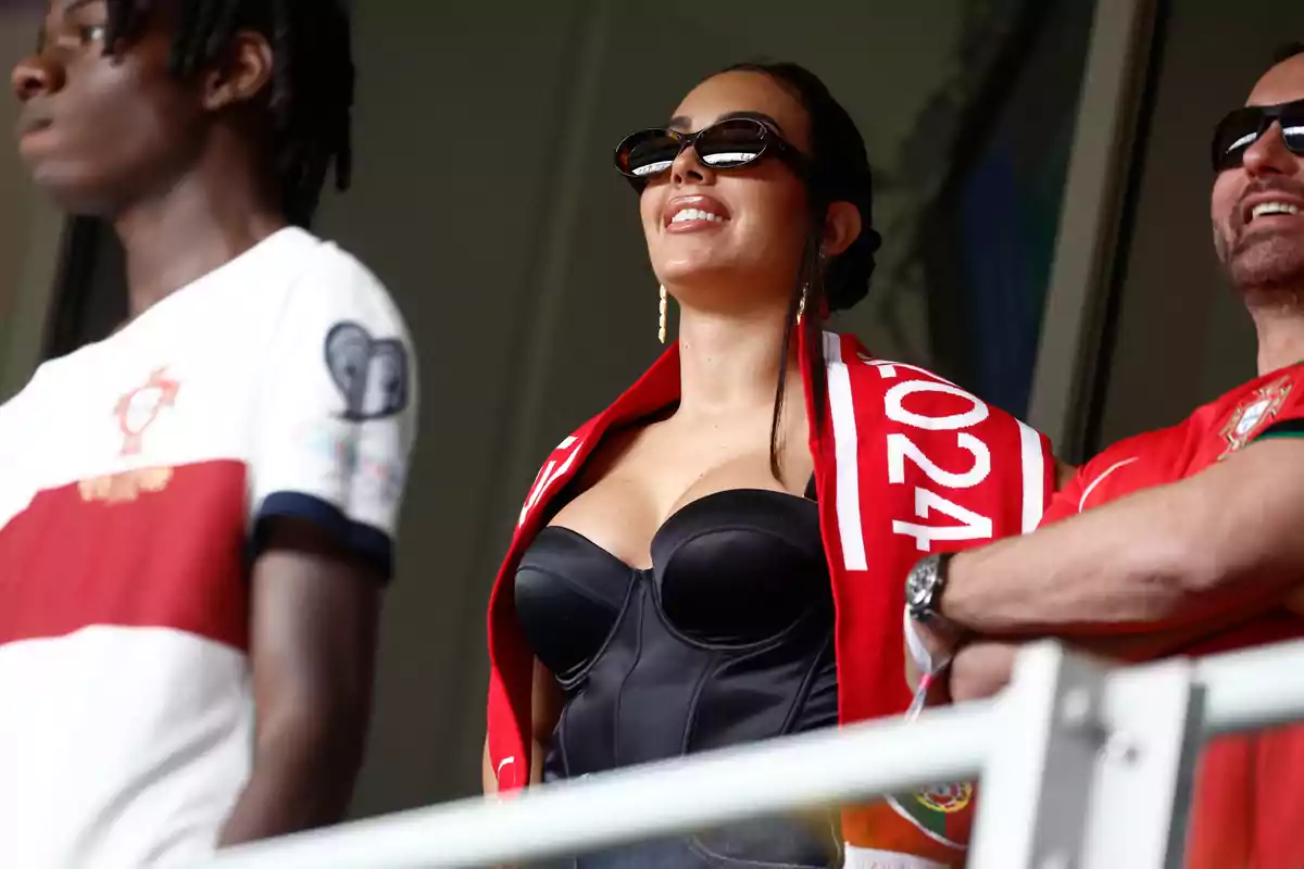 Tres personas con ropa de Portugal observan un evento deportivo, una mujer en el centro lleva gafas de sol y una bufanda roja. Tres personas con ropa de Portugal observan un evento deportivo, una mujer en el centro lleva gafas de sol y una bufanda roja.