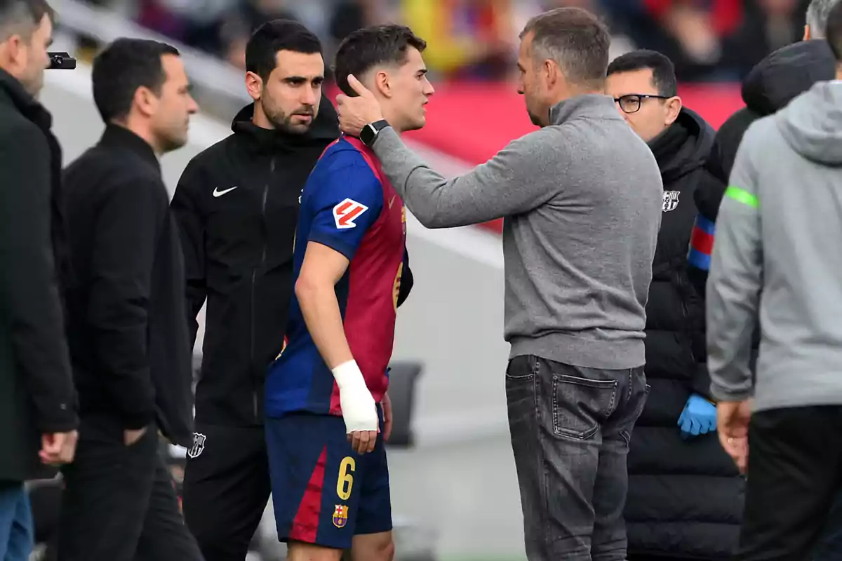 A group of people on a soccer field, one of them in a sports uniform with a bandage on their hand, is being consoled by another person who touches their face.