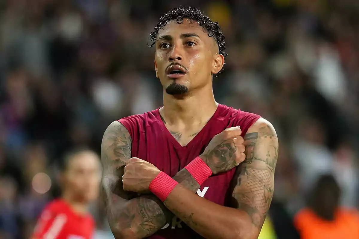 Raphinha con una camiseta sin mangas roja y muñequeras rojas cruza los brazos frente a su pecho en un estadio lleno de espectadores.