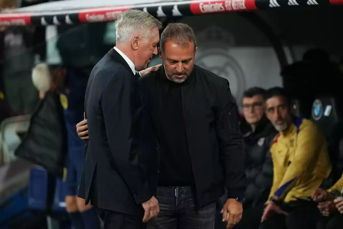 Two men chat closely in a technical area of a stadium while other people watch from the background.