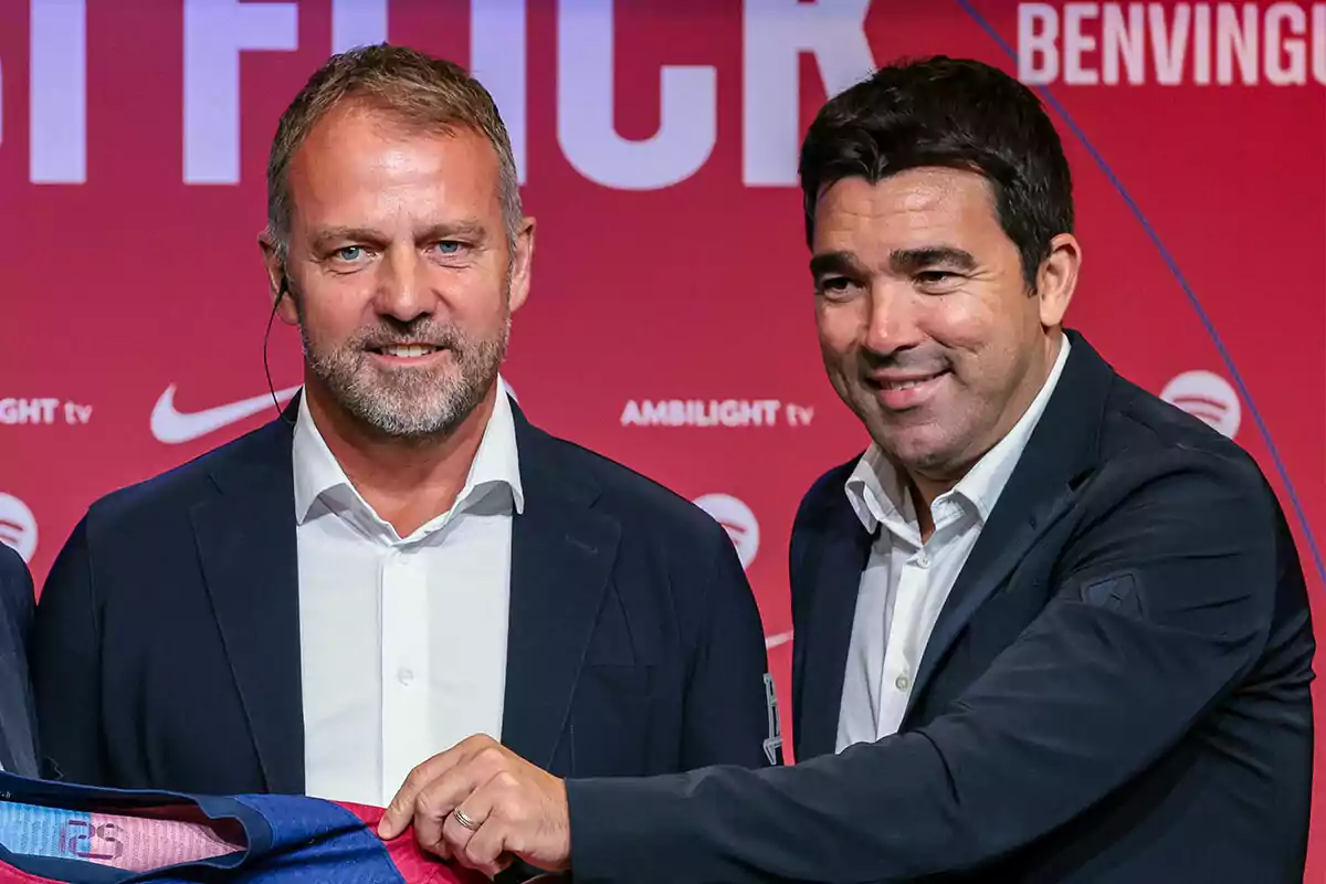 Hansi Flick y Deco posan frente a un fondo rojo con logotipos y texto, uno de ellos sostiene una camiseta deportiva.