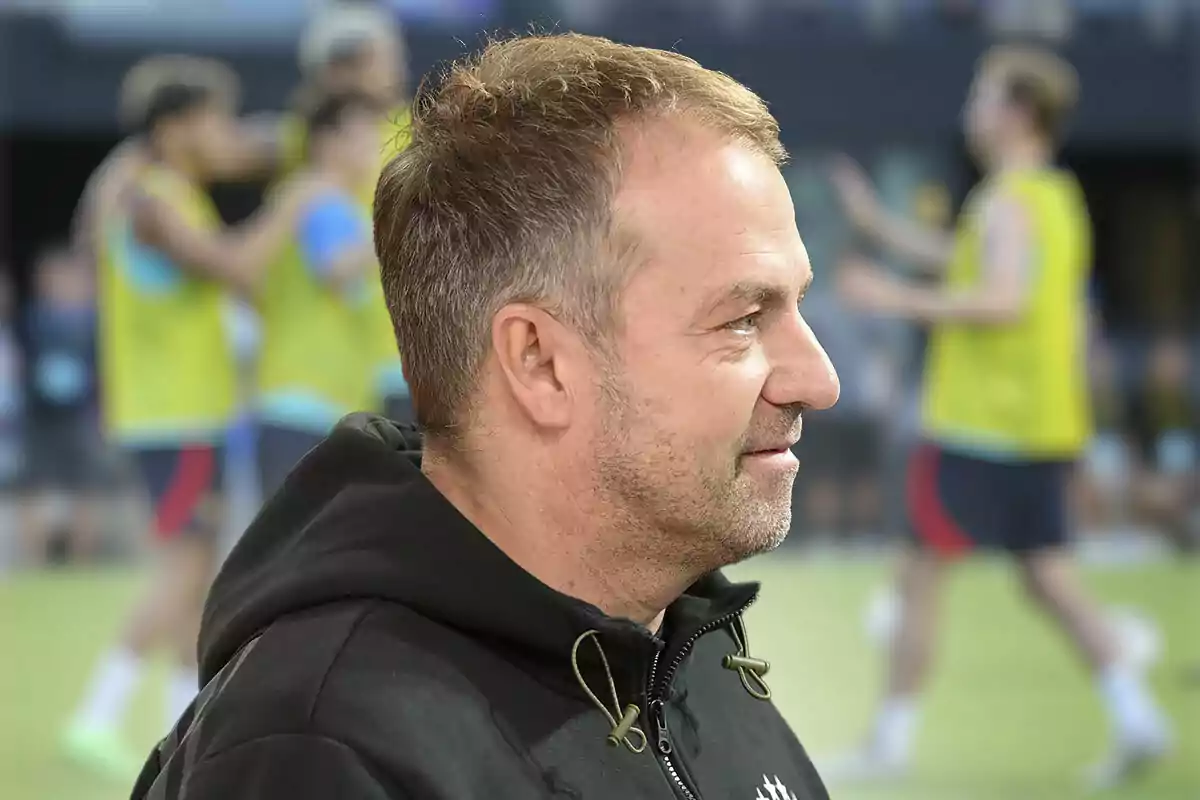 Hansi Flick con una sudadera negra está de perfil en primer plano, mientras que en el fondo se ven varios jugadores del Barça desenfocados con chalecos amarillos en un campo de entrenamiento deportivo.