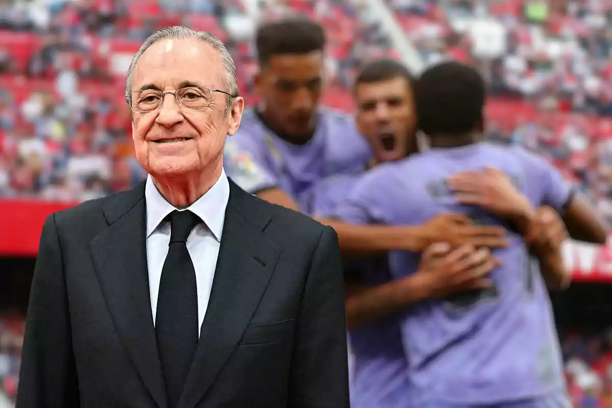 Florentino Pérez con gafas y traje oscuro sonríe frente a un grupo de jugadores del Real Madrid que celebran en el fondo.