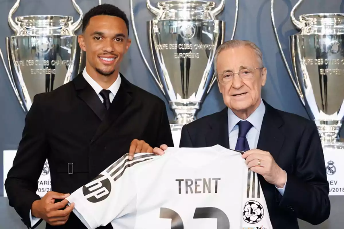 Dos hombres posan sonrientes sosteniendo una camiseta de fútbol blanca con el nombre Trent y el número 22, al fondo se observan trofeos grandes plateados.
