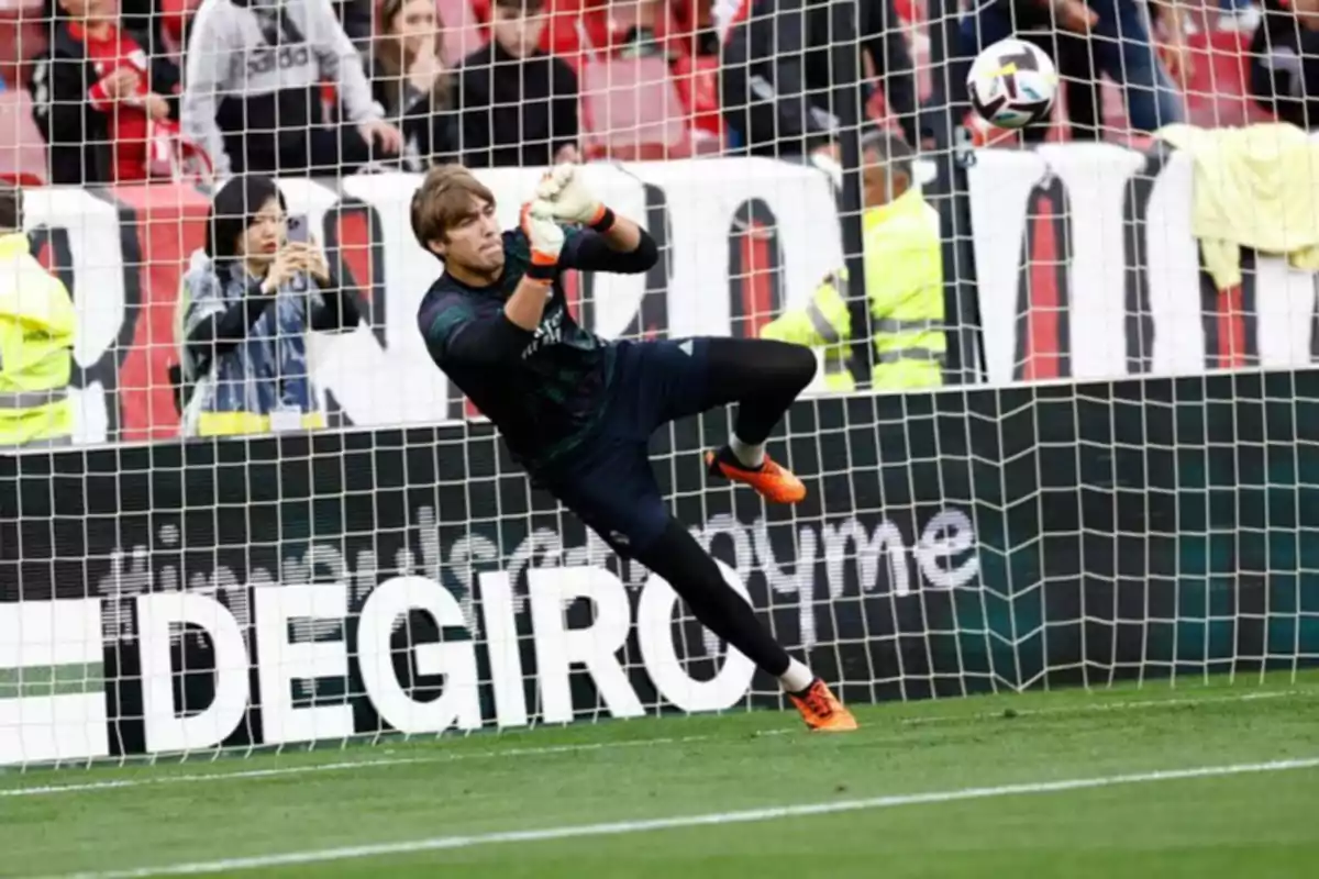 Un portero de fútbol detiene un balón durante un partido con espectadores en el fondo.