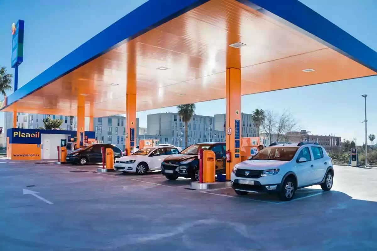 A gas station with several cars parked under an orange and blue roof, with fuel pumps and a building in the background. A gas station with several cars parked under an orange and blue roof, with fuel pumps and a building in the background.