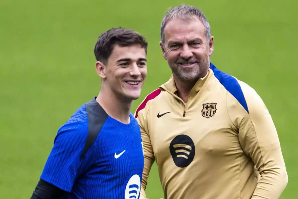 Dos personas sonrientes, una con camiseta azul y otra con chaqueta beige, en un campo de fútbol.