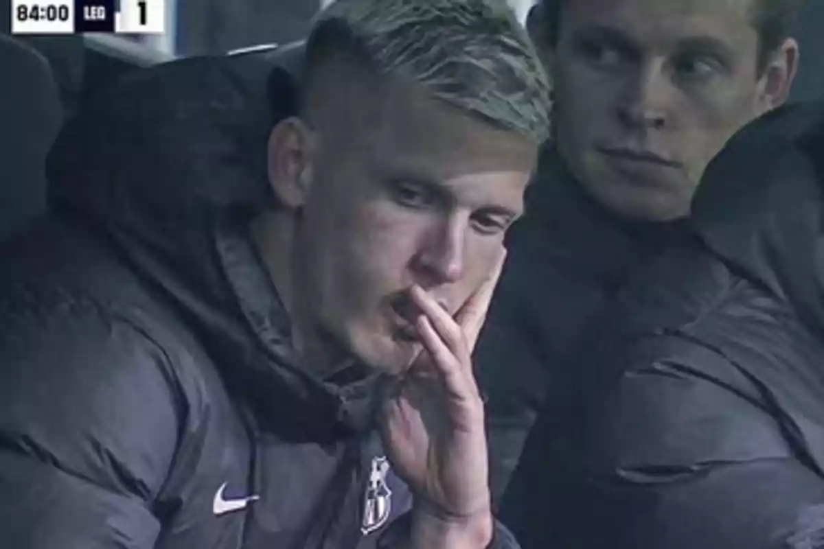 Two people sitting on a bench during a soccer match, one of them looking thoughtful while holding his hand to his mouth.