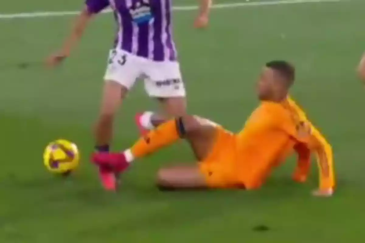 A player in an orange uniform is on the ground while another player in a purple and white uniform approaches the ball.