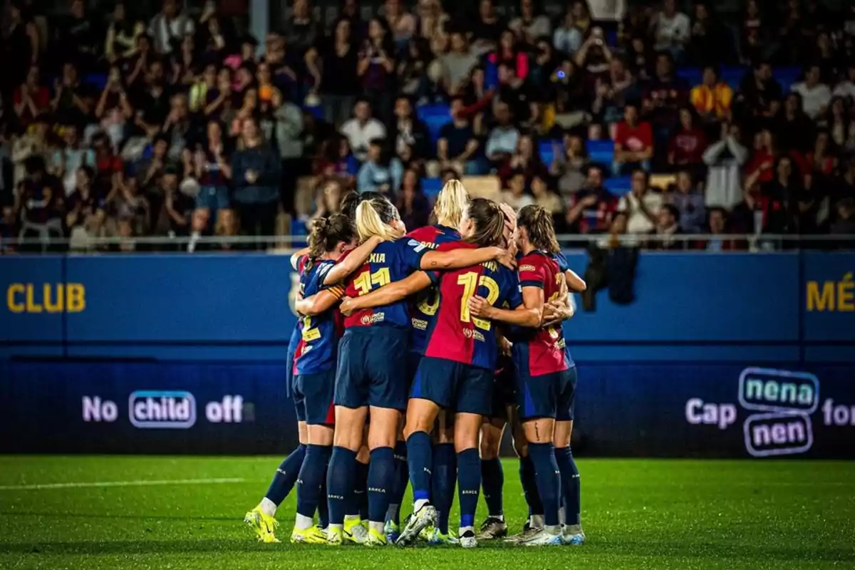 Un grupo de jugadoras de fútbol se abrazan en el campo mientras el público las observa desde las gradas.