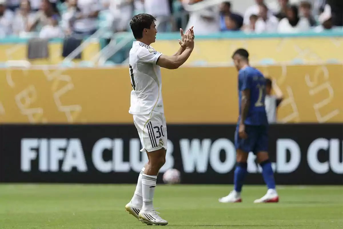 Soccer player in a white uniform clapping on the field during a match with spectators in the stands and an opponent in a blue uniform in the background