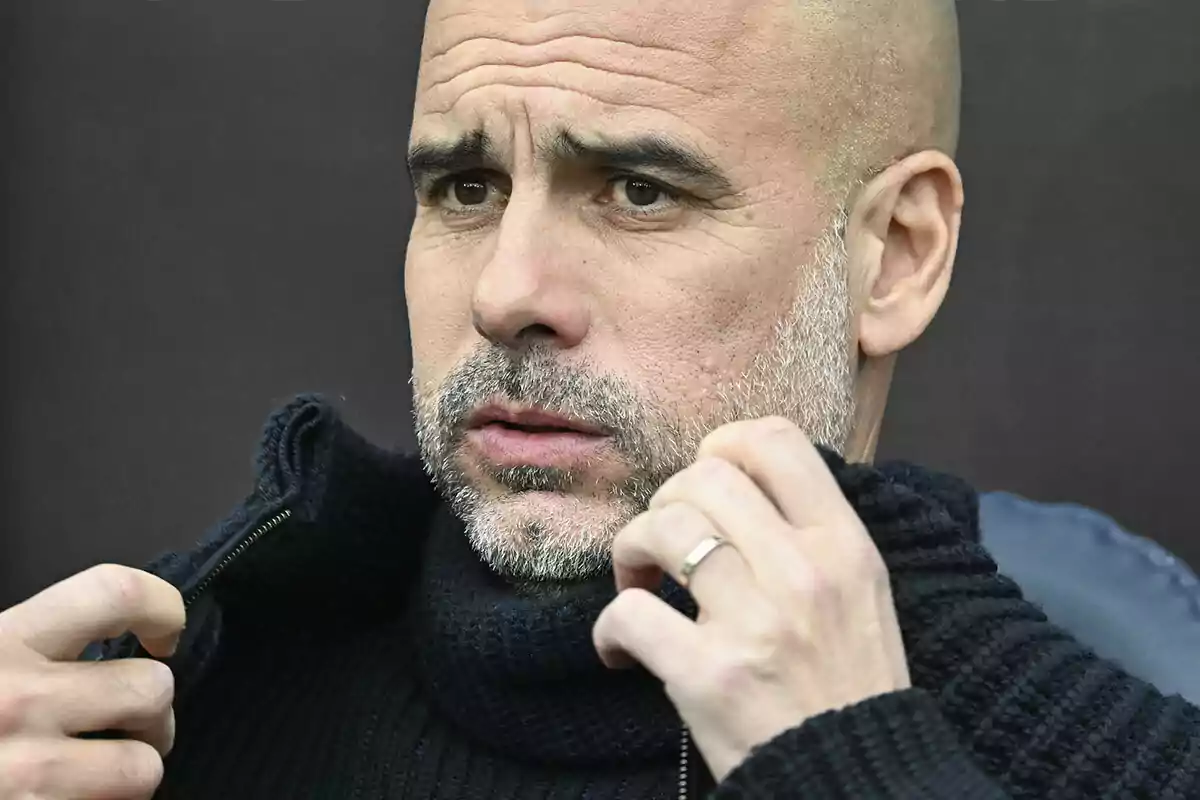 Pep Guardiola con barba canosa y suéter negro ajustándose el cuello del suéter.