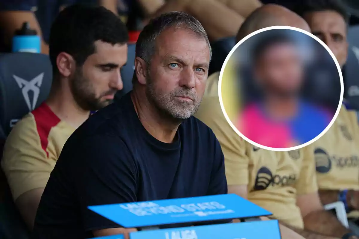 Hansi Flick, vestido con una camiseta oscura, está sentado en un banquillo deportivo junto a otros hombres, uno de los cuales lleva una camiseta beige con detalles rojos.