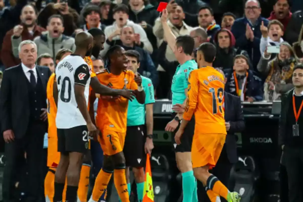 A referee shows a red card to a player in a soccer match as other players and the crowd react.