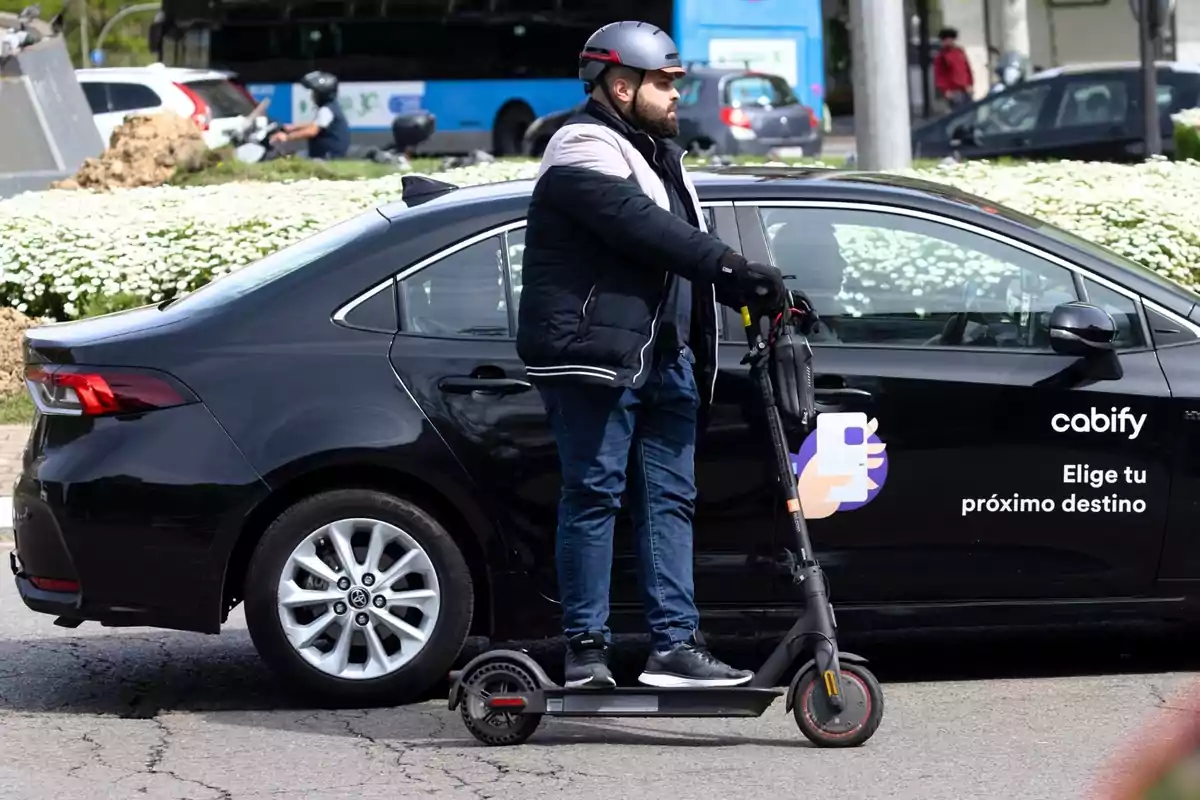 Un hombre con casco y chaqueta oscura monta un patinete eléctrico frente a un coche negro de la empresa Cabify que tiene el eslogan 