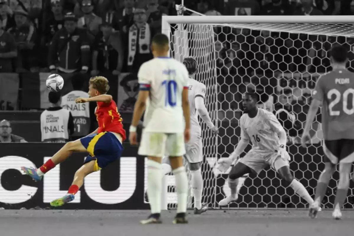 A player in a red and blue uniform strikes the ball in front of the goal while the goalkeeper and other players watch the play in an image where only the red team is in color and the rest are in black and white. A player in a red and blue uniform strikes the ball in front of the goal while the goalkeeper and other players watch the play in an image where only the red team is in color and the rest are in black and white.
