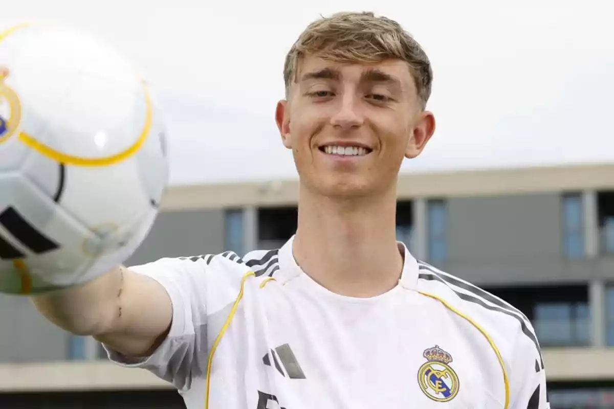 Young man wearing a Real Madrid jersey smiles while holding a soccer ball in front of the camera.